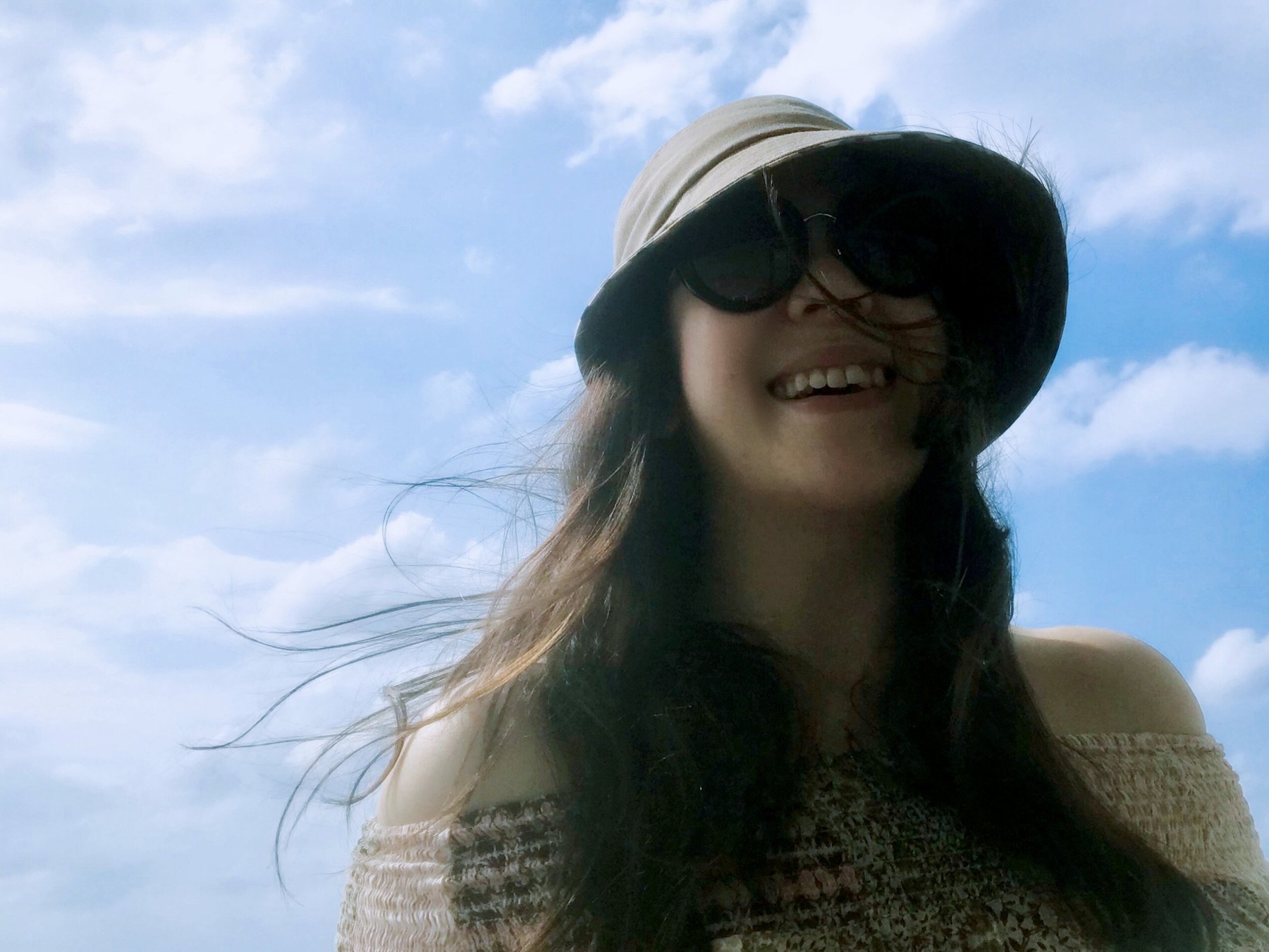 A woman wearing a sun hat and sunglasses, smiling against a background of blue sky and clouds.