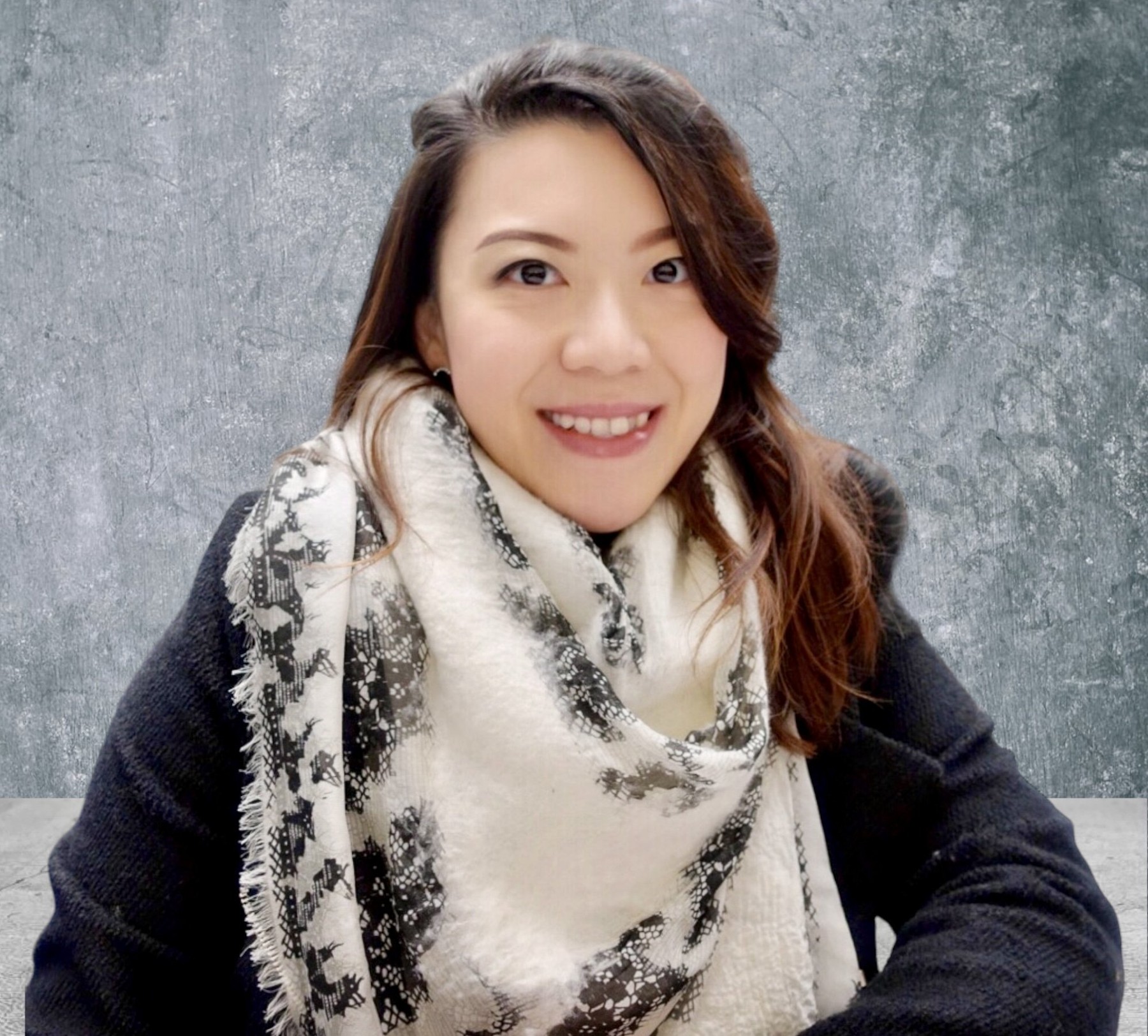 Smiling woman with shoulder-length wavy hair, wearing a black sweater and a white scarf with black patterns, sitting in front of a textured gray background.