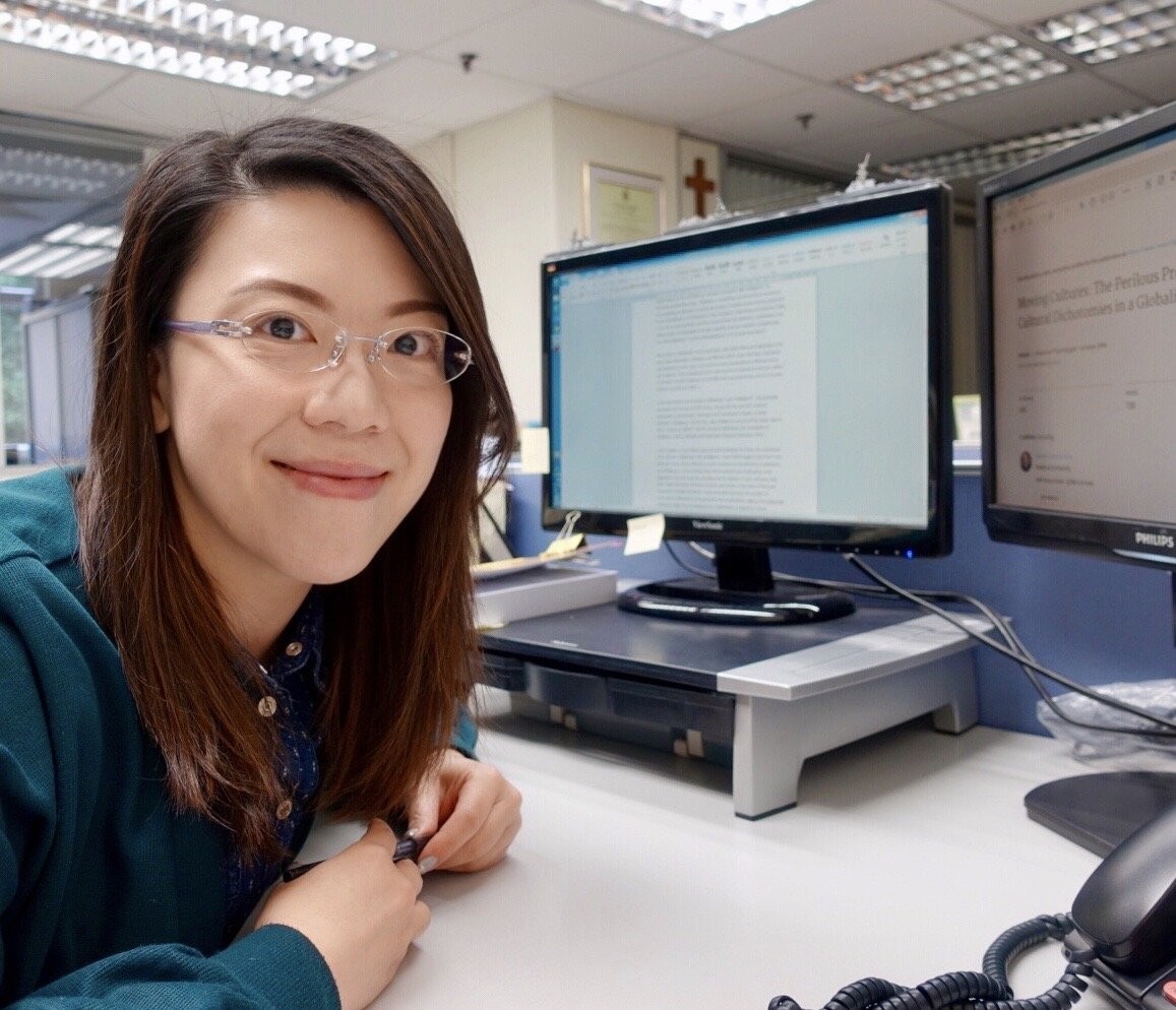 A woman with glasses sitting at a desk in an office, smiling at the camera, with two computer monitors in front of her displaying text documents.