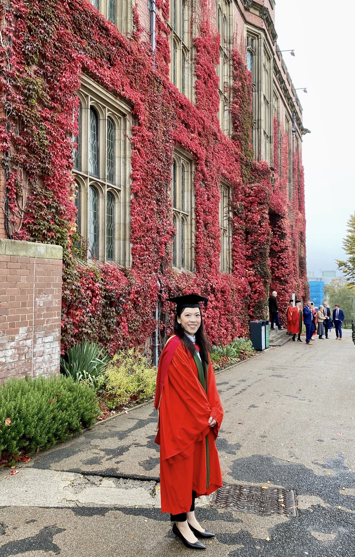 Woman in graduation gown and cap standing outdoors in front of a building covered in red and green ivy, with other graduates and people in the background.