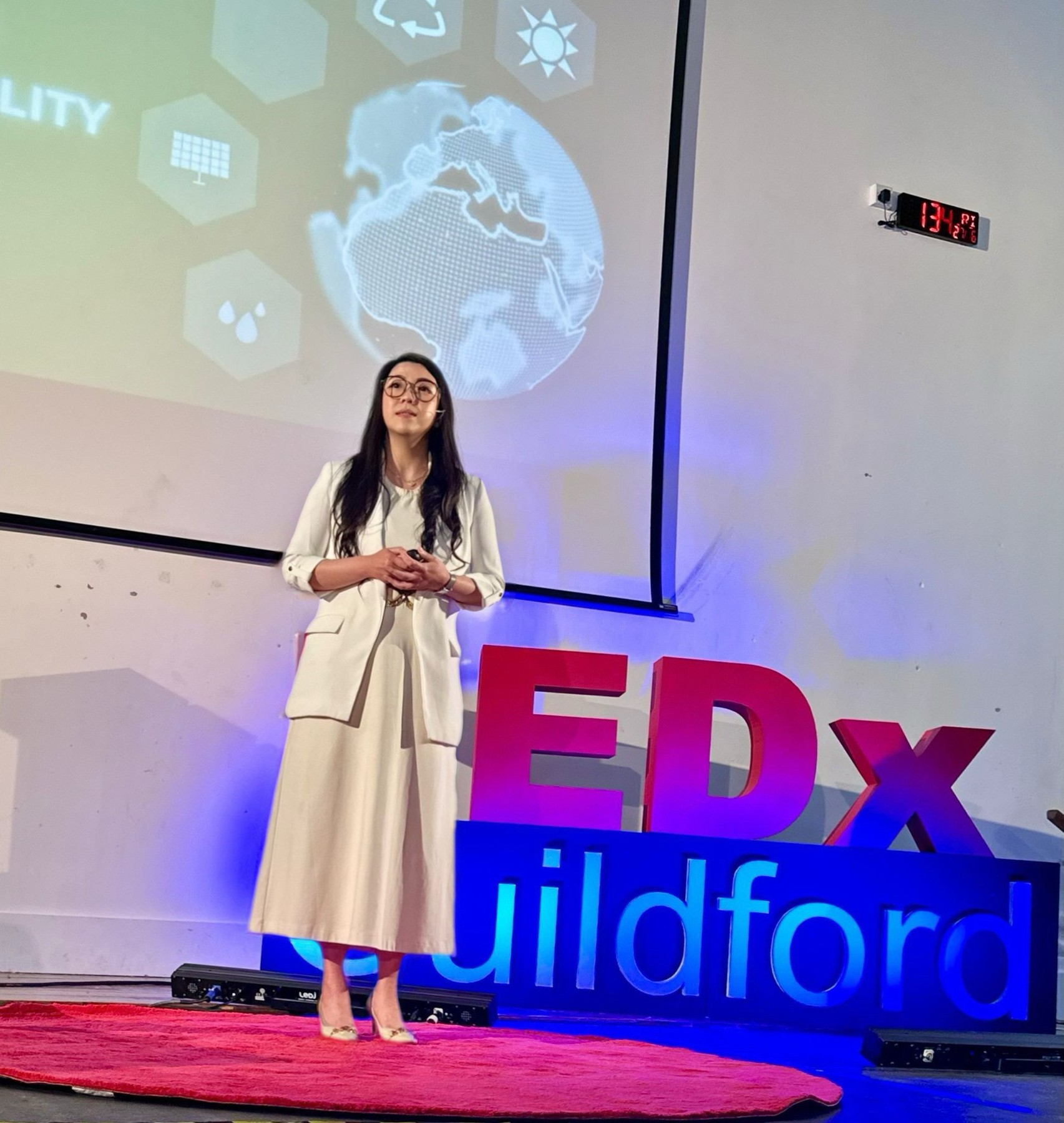 A woman in a white pantsuit standing on a red stage with large colorful 3D letters spelling 'EDX' and 'wildford'. She is giving a presentation at a TEDx event, with a large screen showing a globe and eco-related icons behind her.