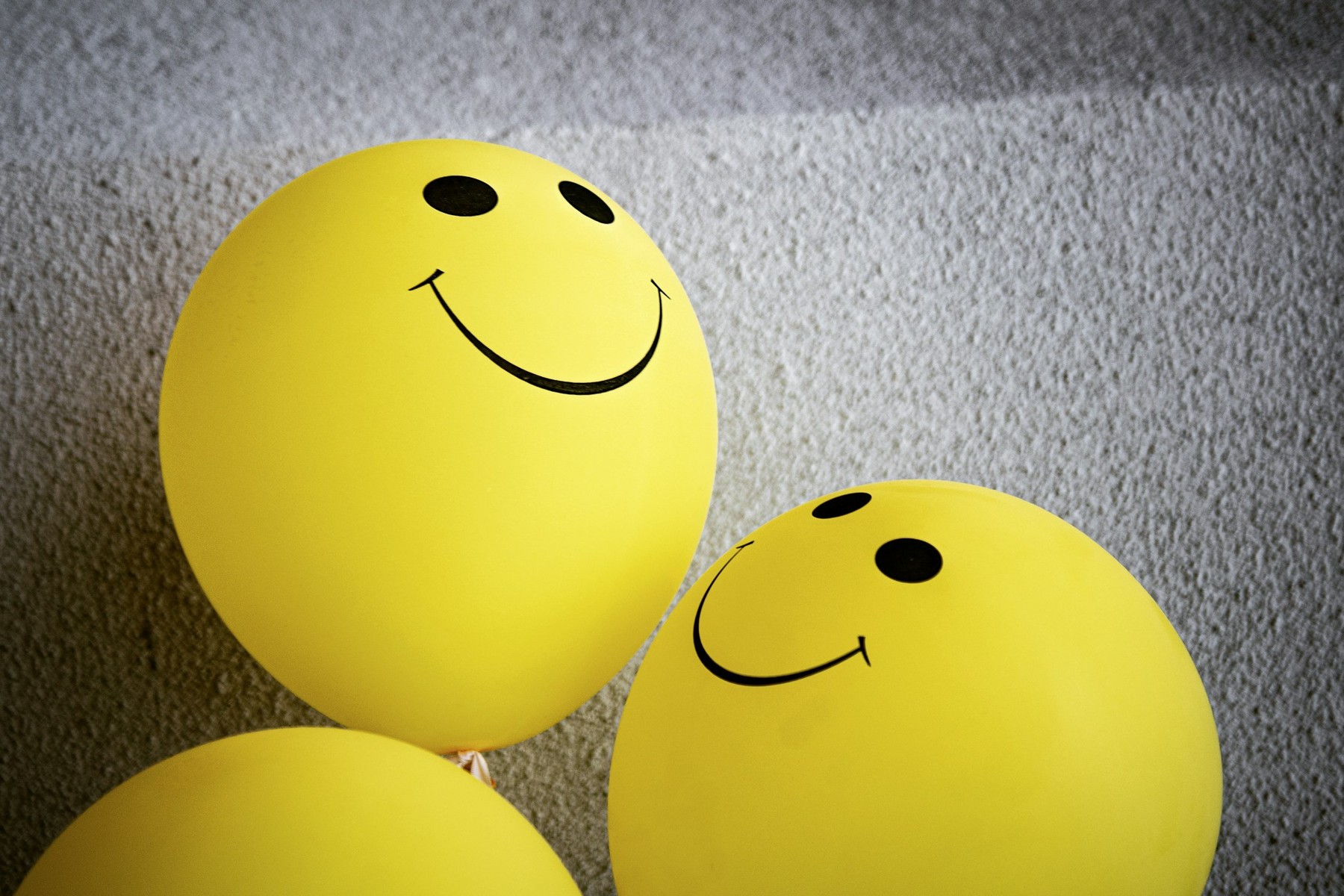 Three yellow balloons with smiley face stickers on a textured wall.
