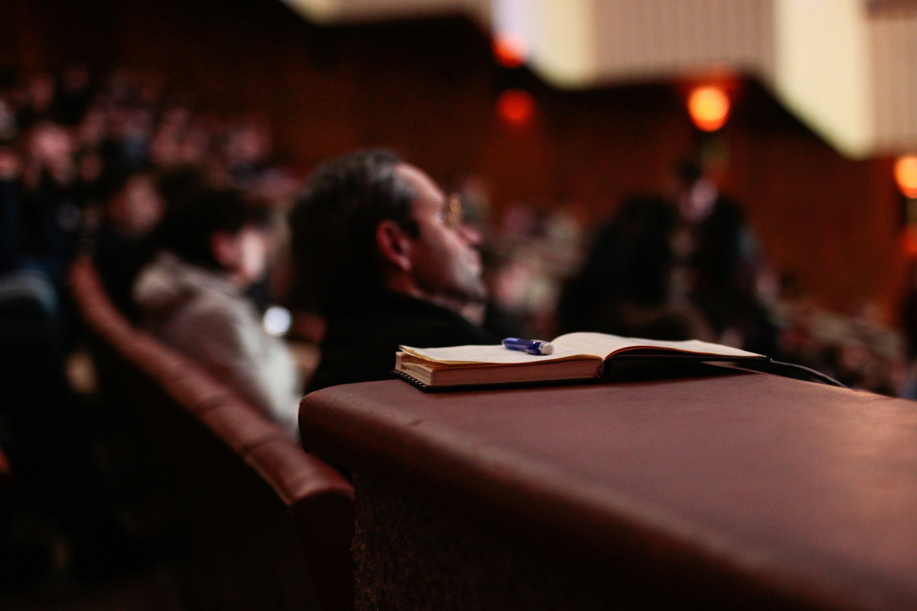 A book and a pen resting on a table in a dimly lit conference or seminar room with people seated and listening.