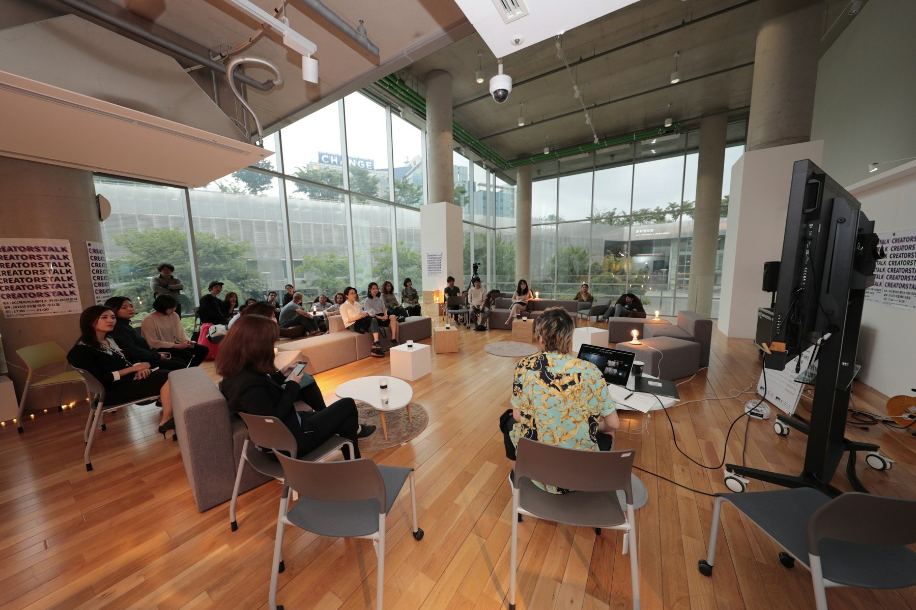 A seminar or workshop taking place in a modern, spacious room with large glass windows, wooden floors, and chairs arranged in a semi-circle. A presenter is seated at a table with a laptop, facing the audience. People are seated listening attentively, with some using their phones. There are posters on the wall, and a videoconferencing device is visible.