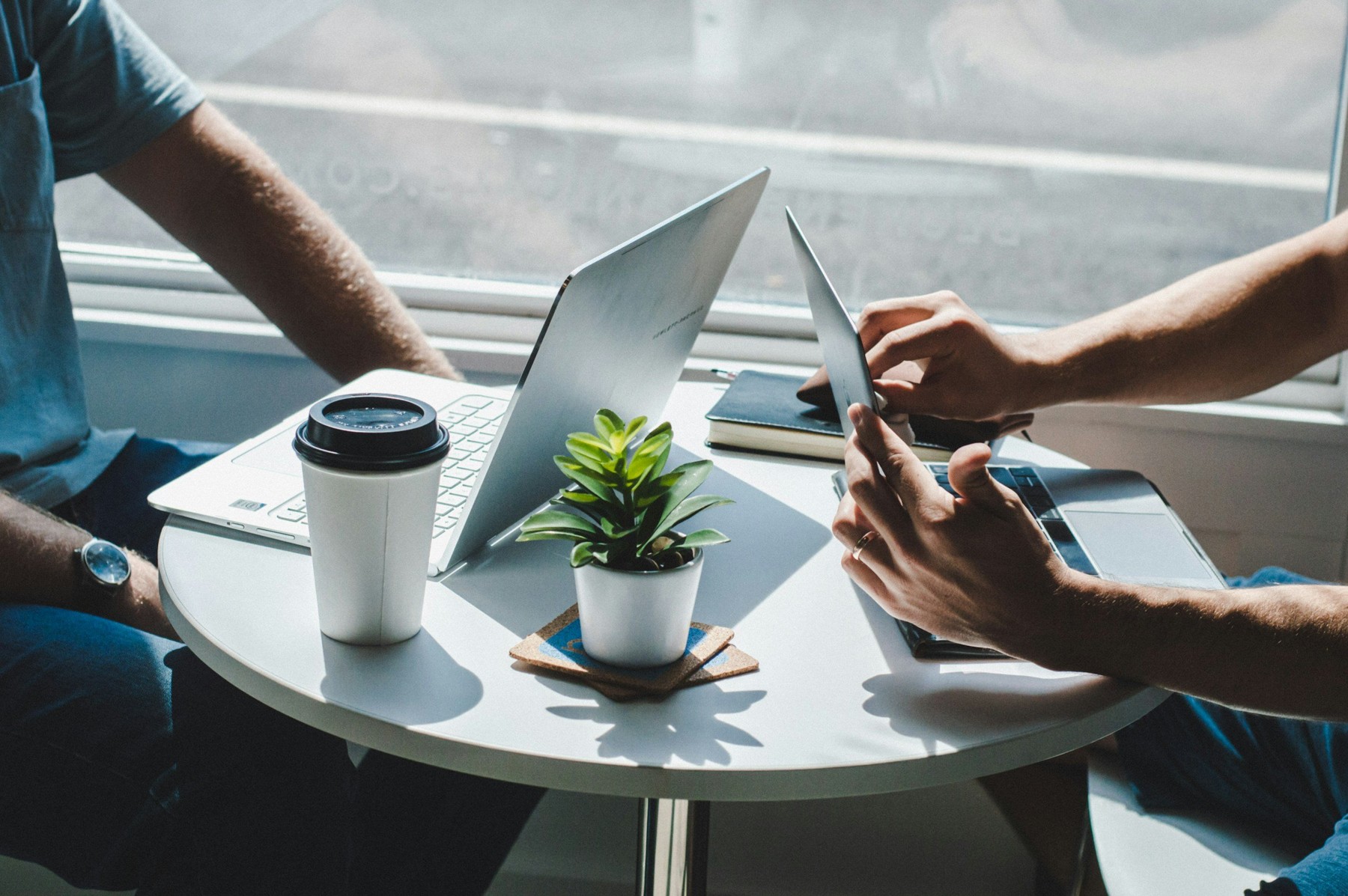 Two people working at a white table with a laptop, tablet, coffee cup, small potted plant, and notebook, near a window with natural light.